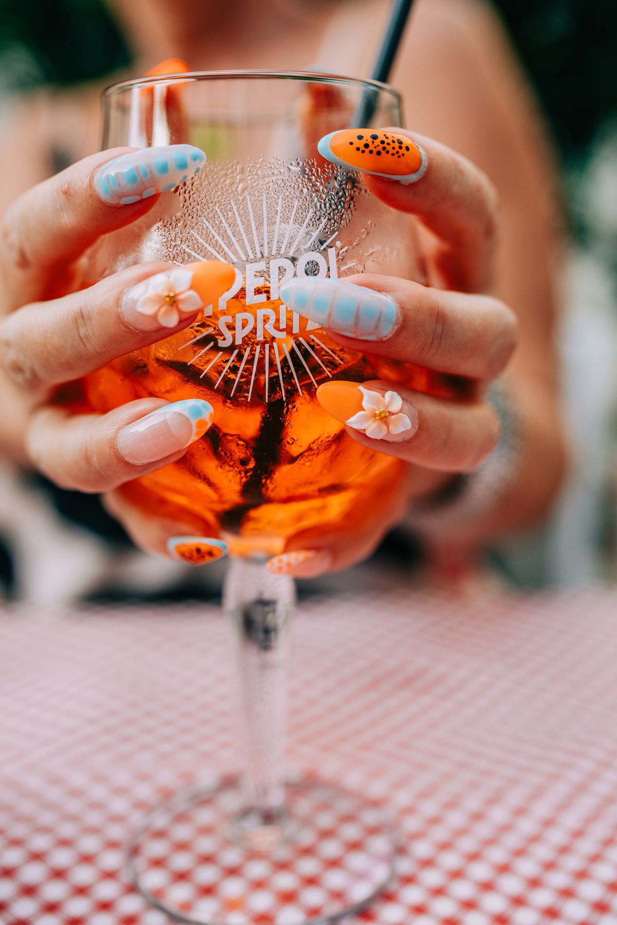 Close-up of hands with colorful nail art holding an Aperol spritz on a checkered tablecloth.
