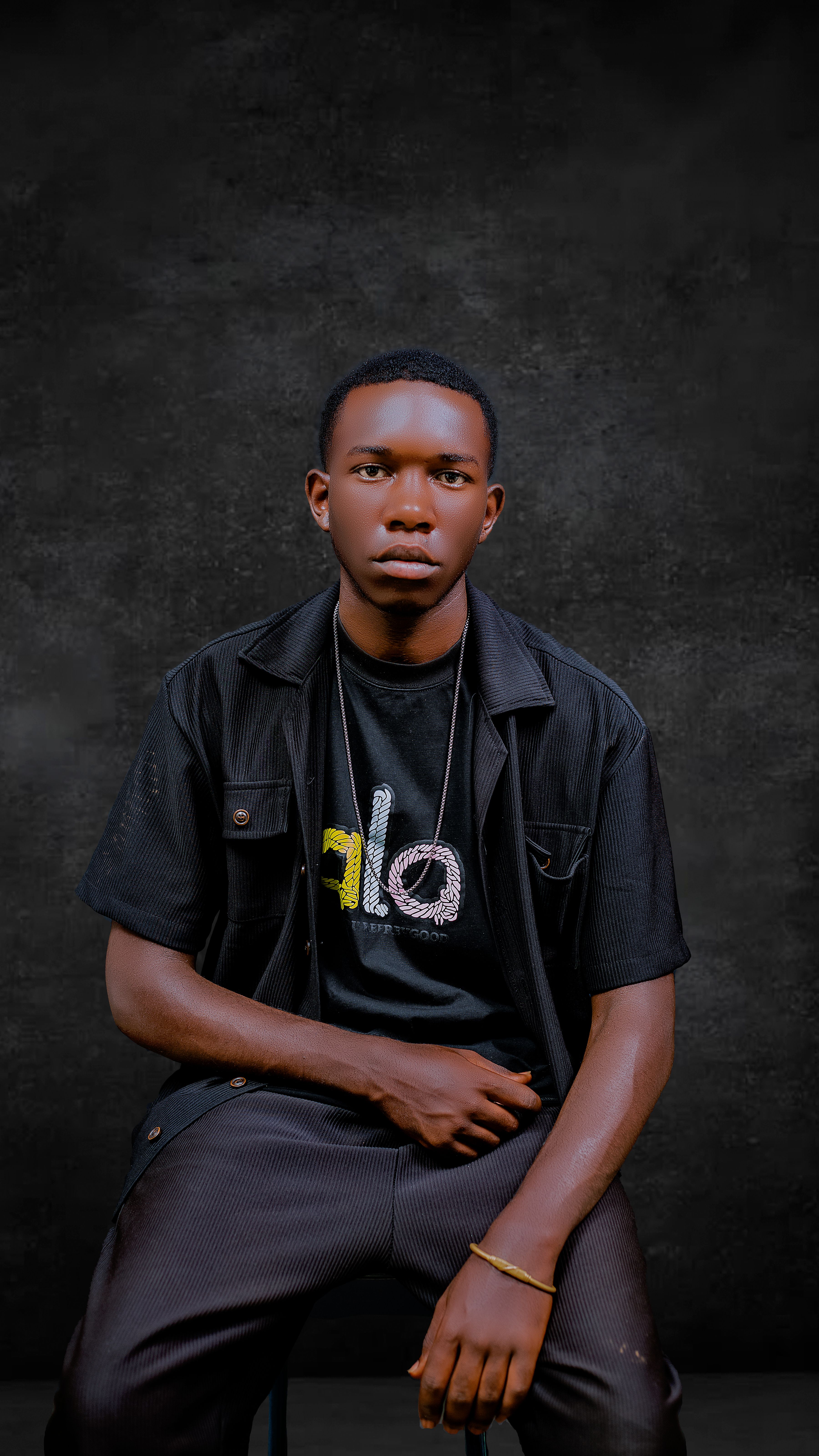 Portrait of a young man in a stylish outfit, seated with a dark textured backdrop.