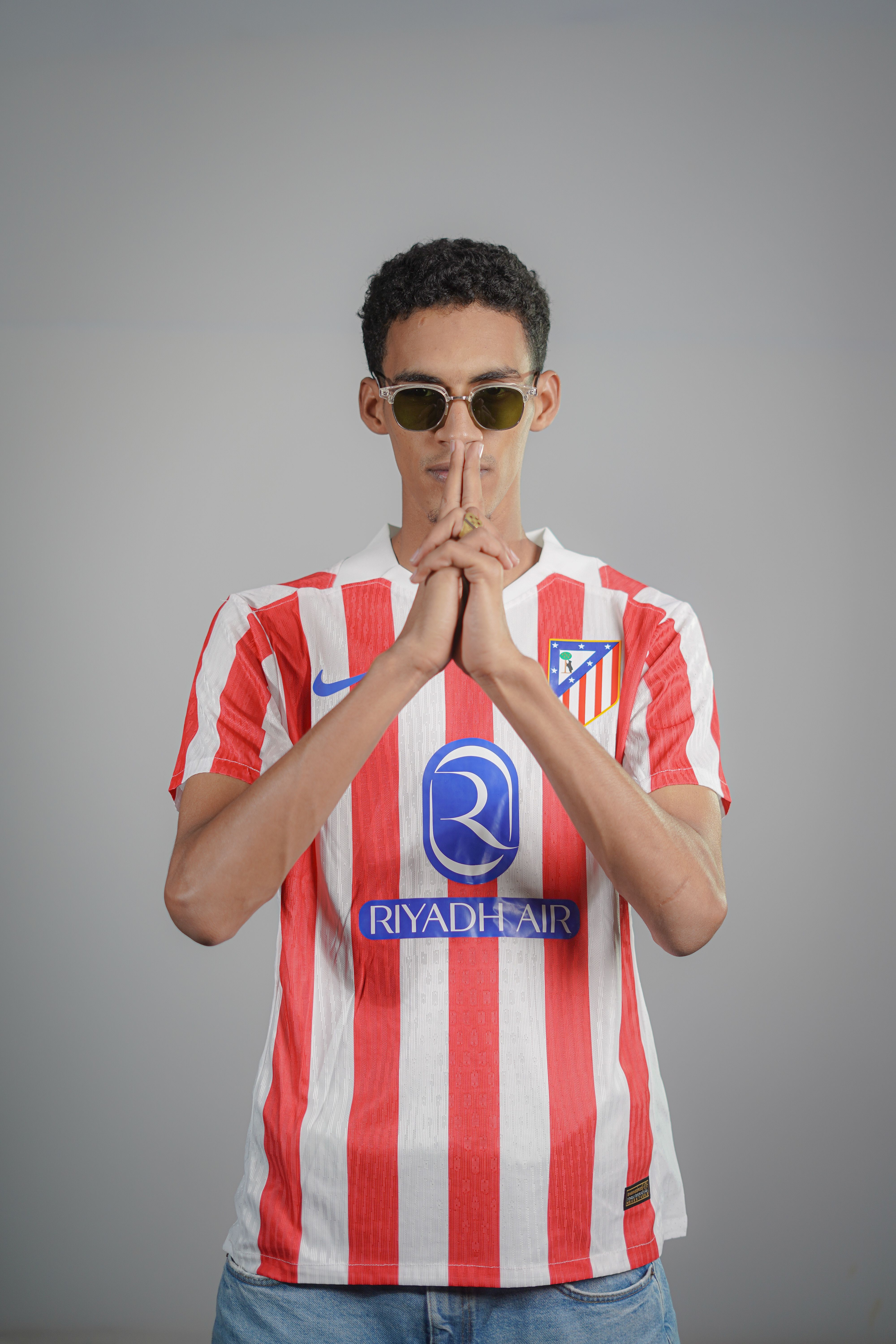 A young man wearing sunglasses and a soccer jersey posing confidently indoors.