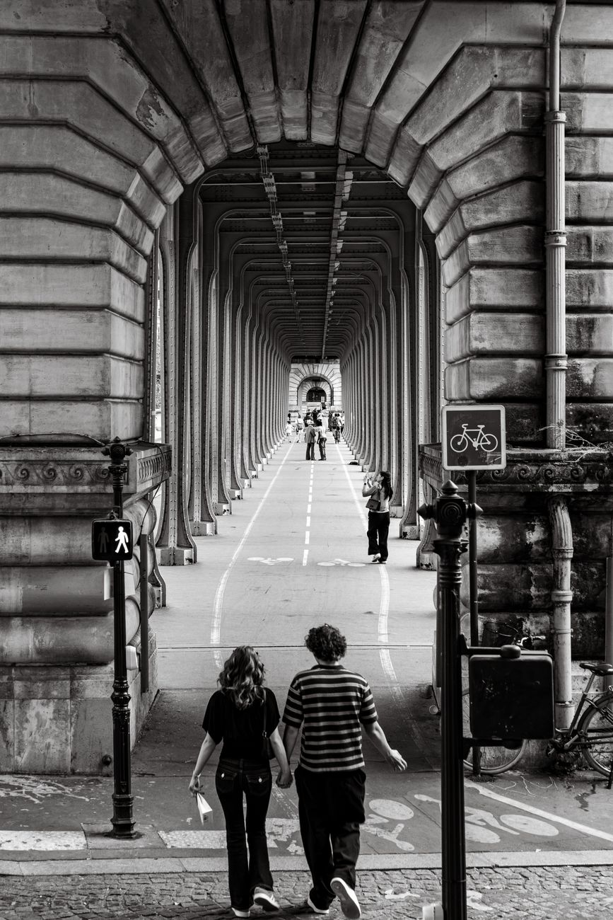 A couple holding hands walks through a historic bridge archway in Paris, France.