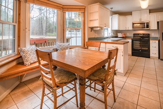 Bright kitchen with wooden dining table, chairs, and large windows offering natural light.