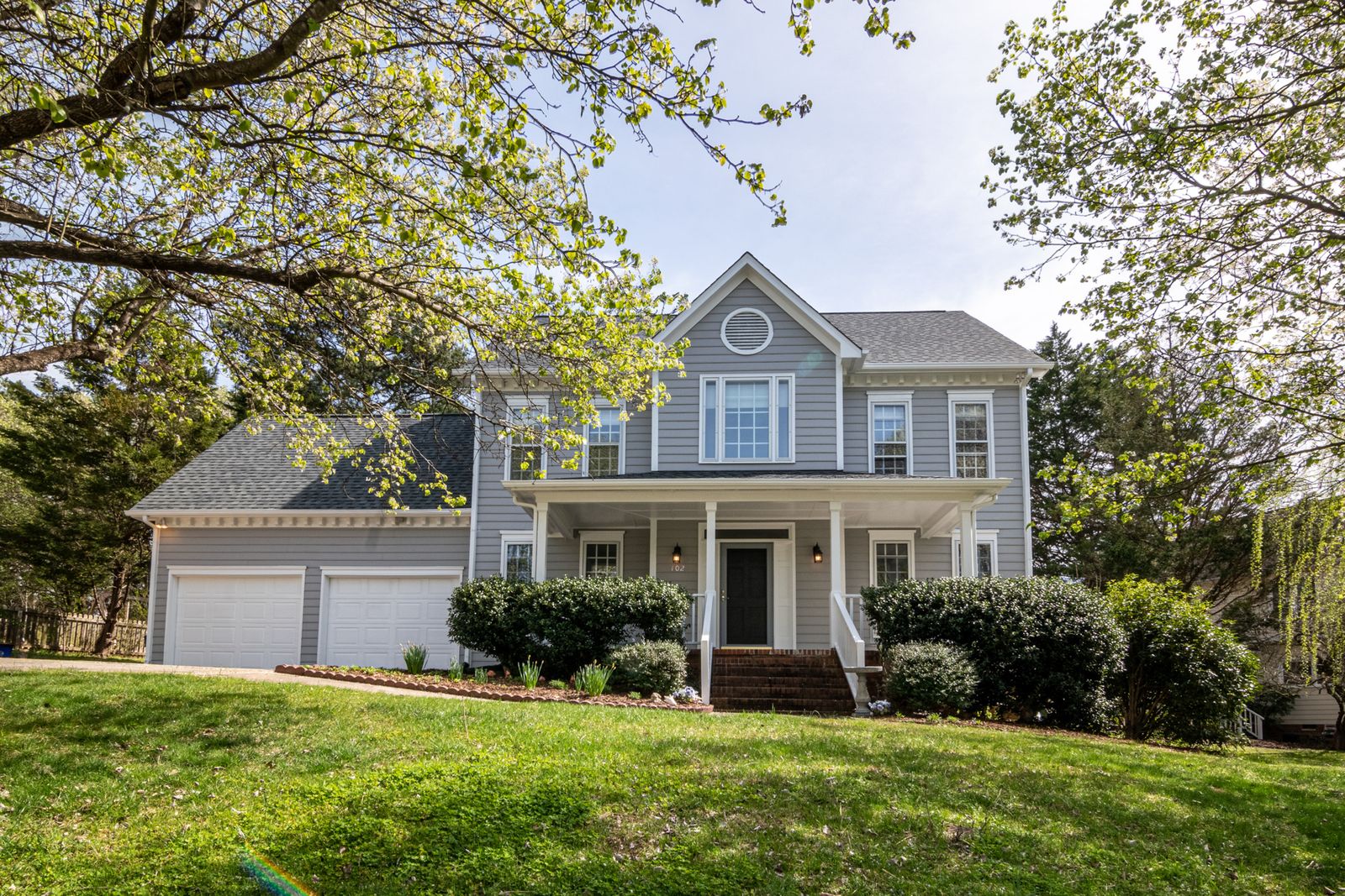 Two-story home with gray-blue siding, a covered front porch, attached garage, and mature spring tree canopy, Carriage Hill, Naperville IL
