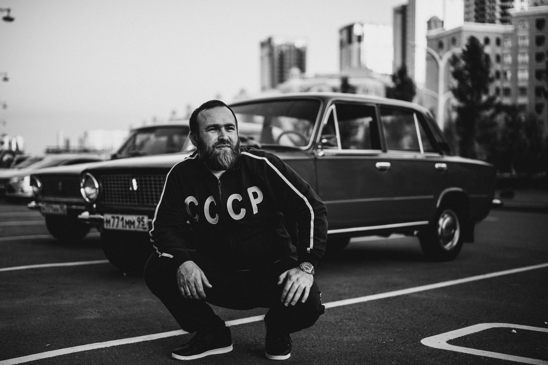 Man in USSR jacket poses by Soviet-era car in urban Grozny. Classic meets modern in black and white.