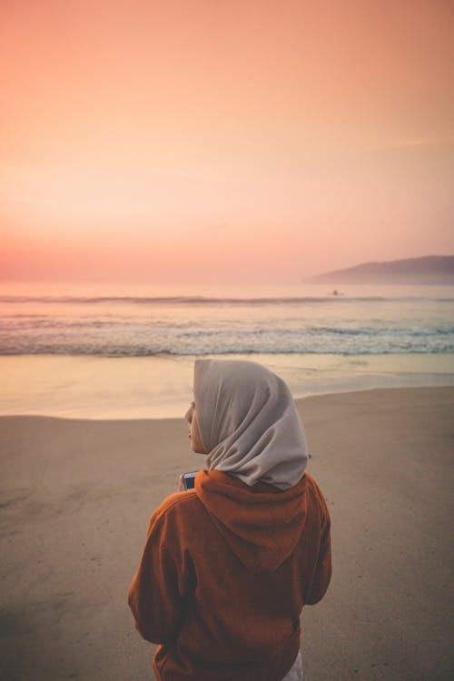 갈색 모래 해변에 서있는 Headscarf를 착용하는 사람