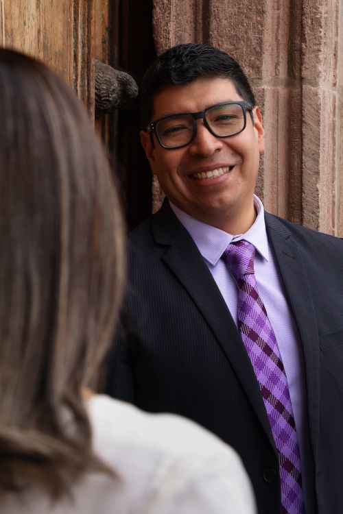 Free Portrait of a man in a suit and purple tie smiling warmly outdoors near a rustic wooden door. Stock Photo