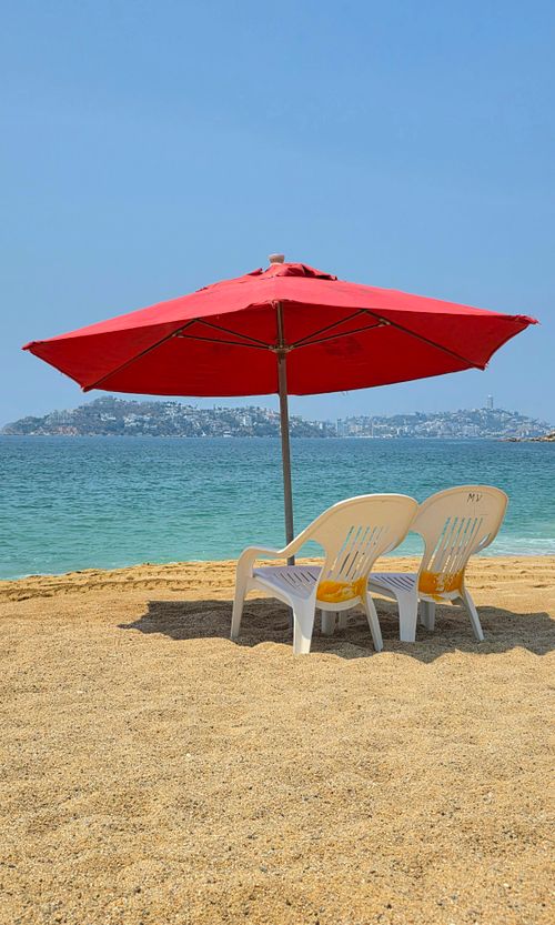 Free Serene beach setting with chairs and a red umbrella by the blue ocean. Stock Photo