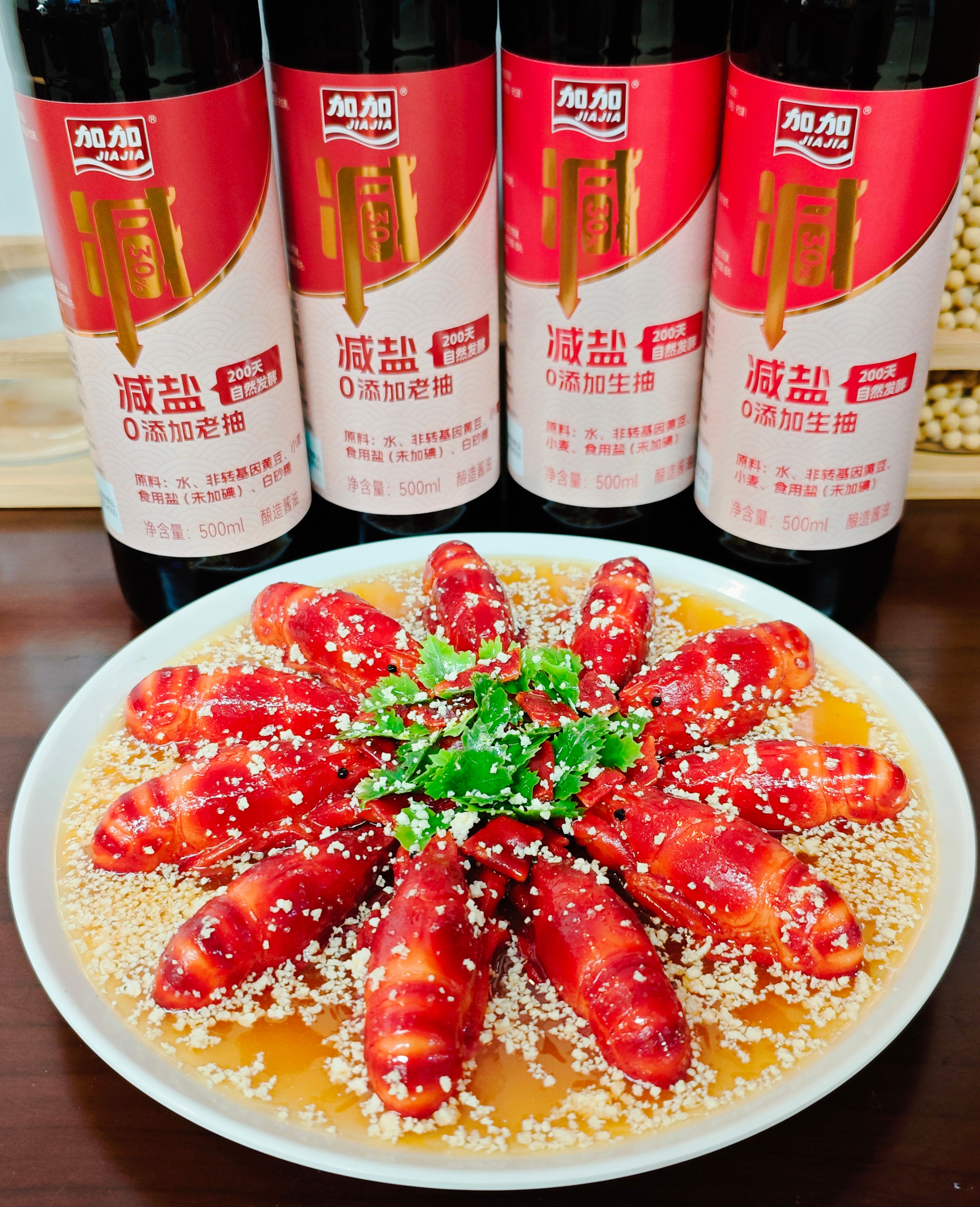 A small glass bottle of dark, glossy homemade soy-free soy sauce substitute being poured over a spoon, with fresh ginger, garlic cloves, and a piece of dried kombu on the counter beside it.