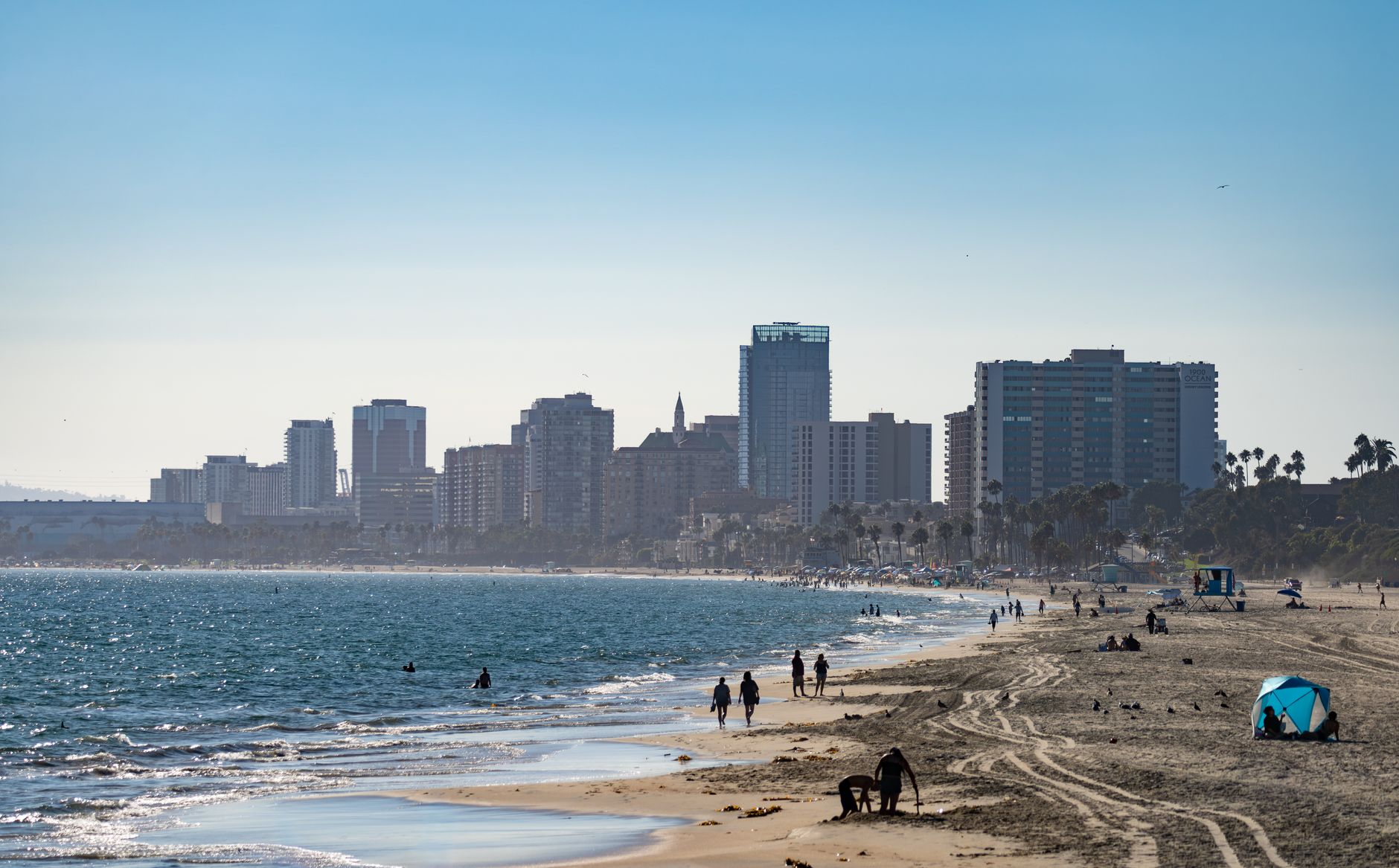 People enjoying a sunny day along Long Beach shore with city skyline in the background.