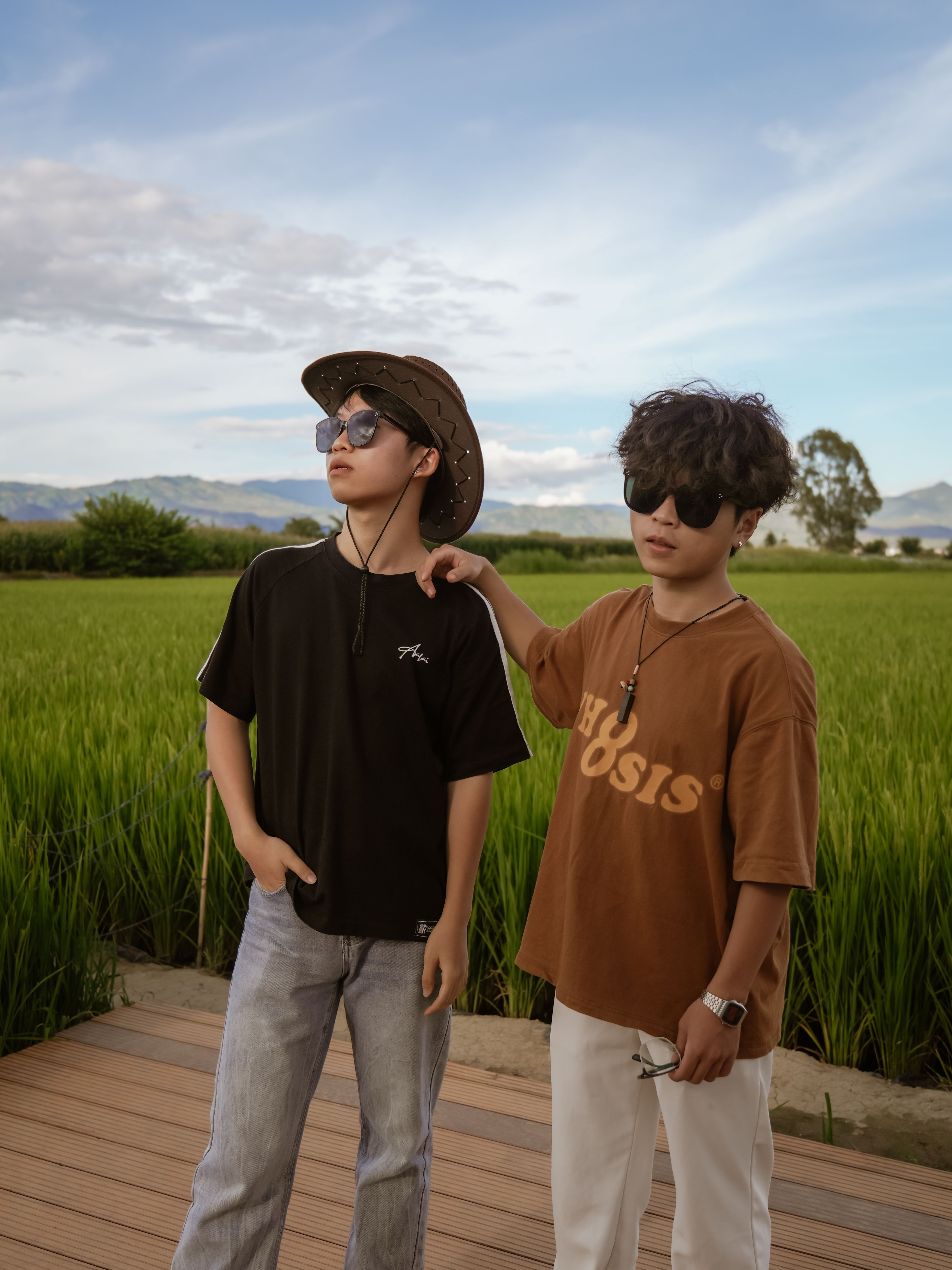 Two friends in trendy outfits and sunglasses pose confidently in a verdant field under a clear blue sky.