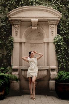A woman in a white dress poses elegantly by a historical stone fountain in Mexico City.