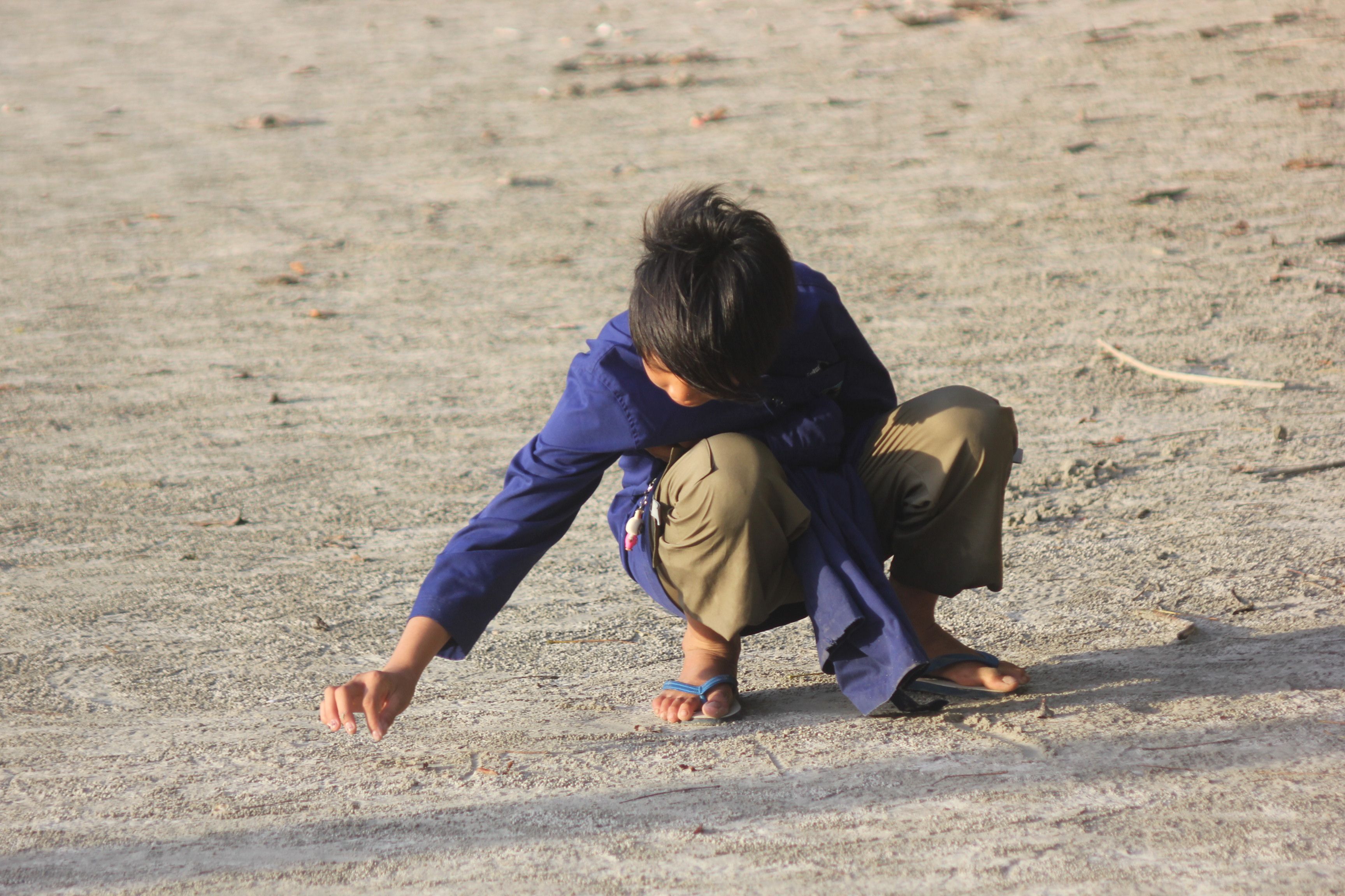 A young child in casual clothes squats on a sandy ground, picking something up in daylight.