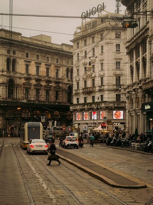 Free A bustling street scene in Milan featuring a tram, classic architecture, and shopping signs with pedestrians crossing. Stock Photo
