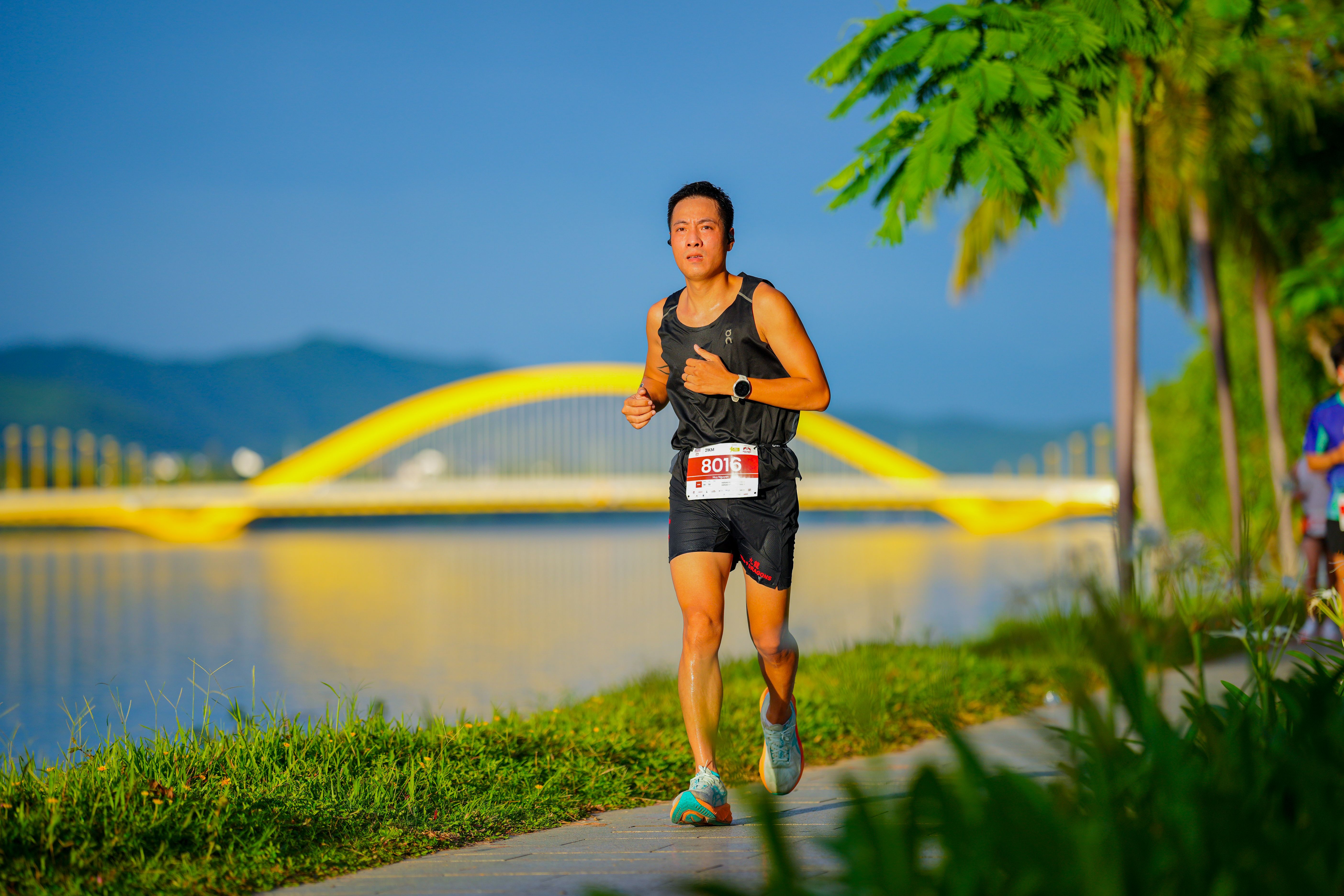 Athlete running along a picturesque riverside with bridge in view.