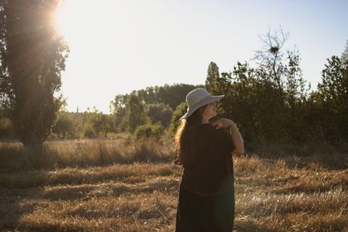 Free A woman in a sun hat enjoys a rural landscape during sunset, creating a serene and warm atmosphere. Stock Photo