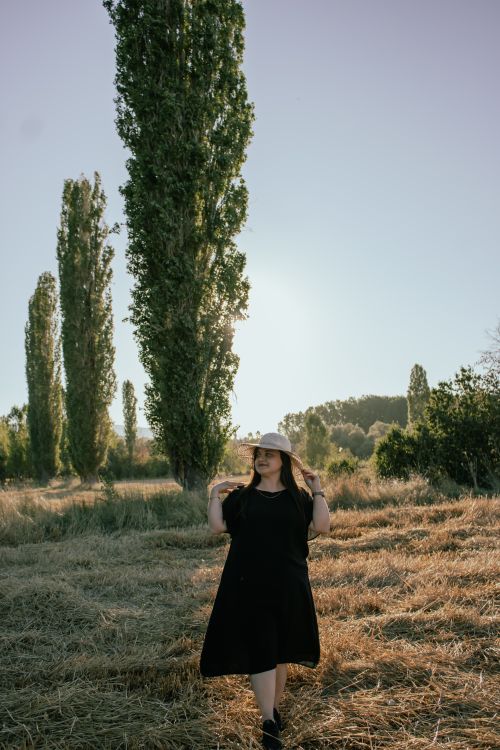 Free A woman in a black dress and hat walking through a field with tall trees during sunrise, embodying nature and serenity. Stock Photo