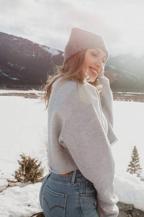 Woman in Gray Sweater and Blue Denim Jeans Standing on Snow Covered Ground