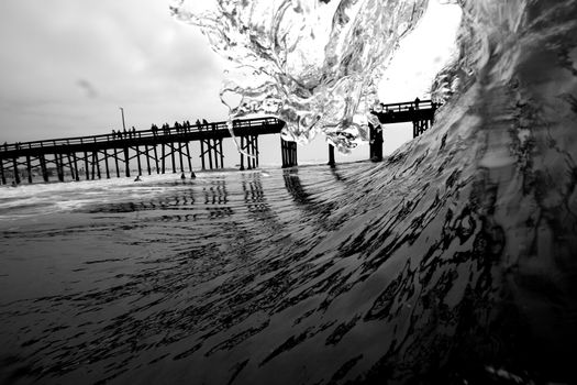Captivating black and white shot of ocean wave breaking near a pier with silhouettes of people.