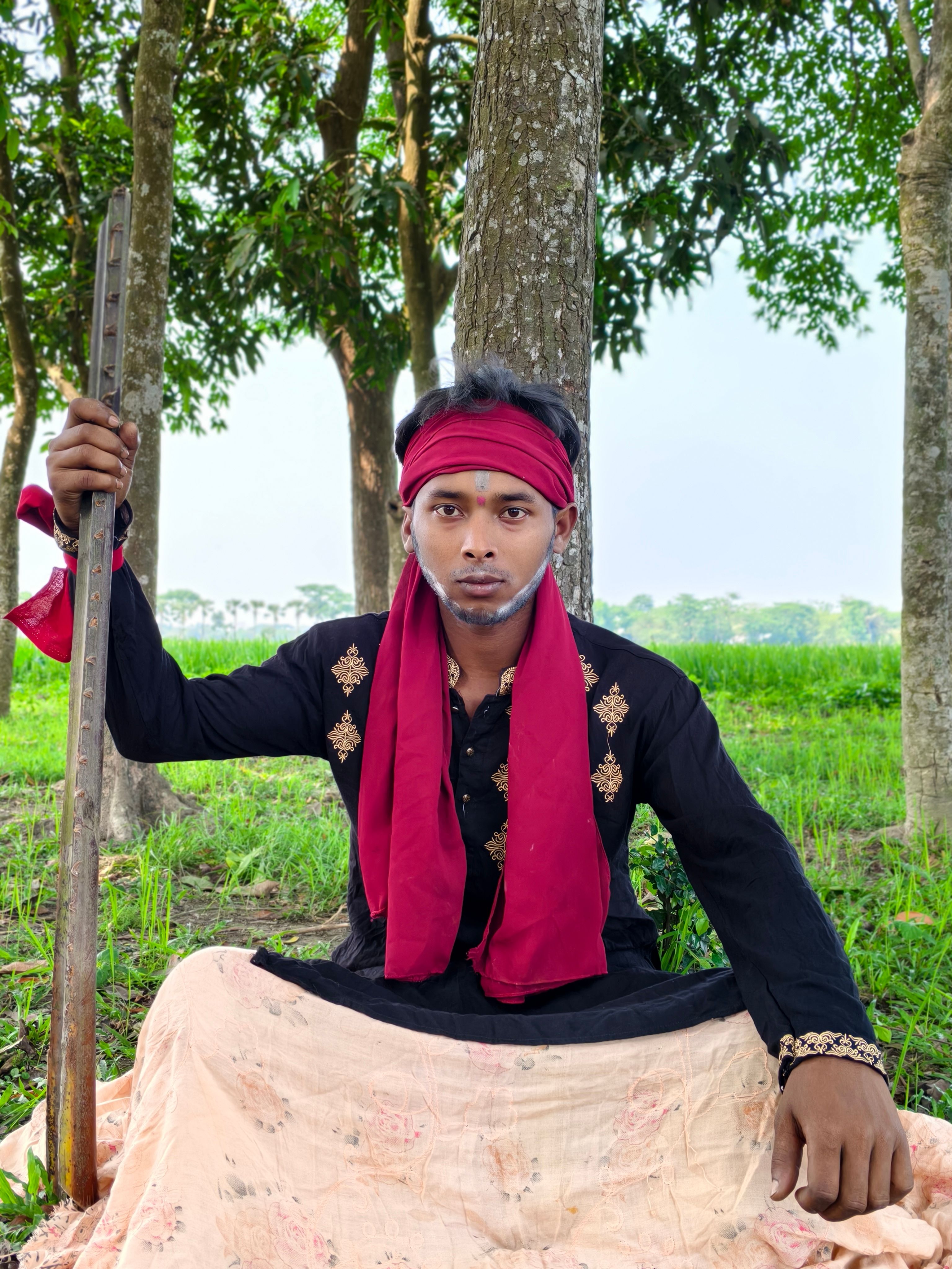 A man in traditional attire poses with a stick outdoors amongst trees and greenery.