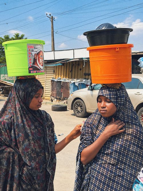 Free Two women carry colorful buckets on their heads in a vivid street market setting. Stock Photo