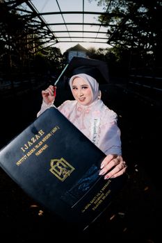 A young woman celebrating graduation, proudly holding her diploma in a cap and gown. - Fotografer Sidoarjo