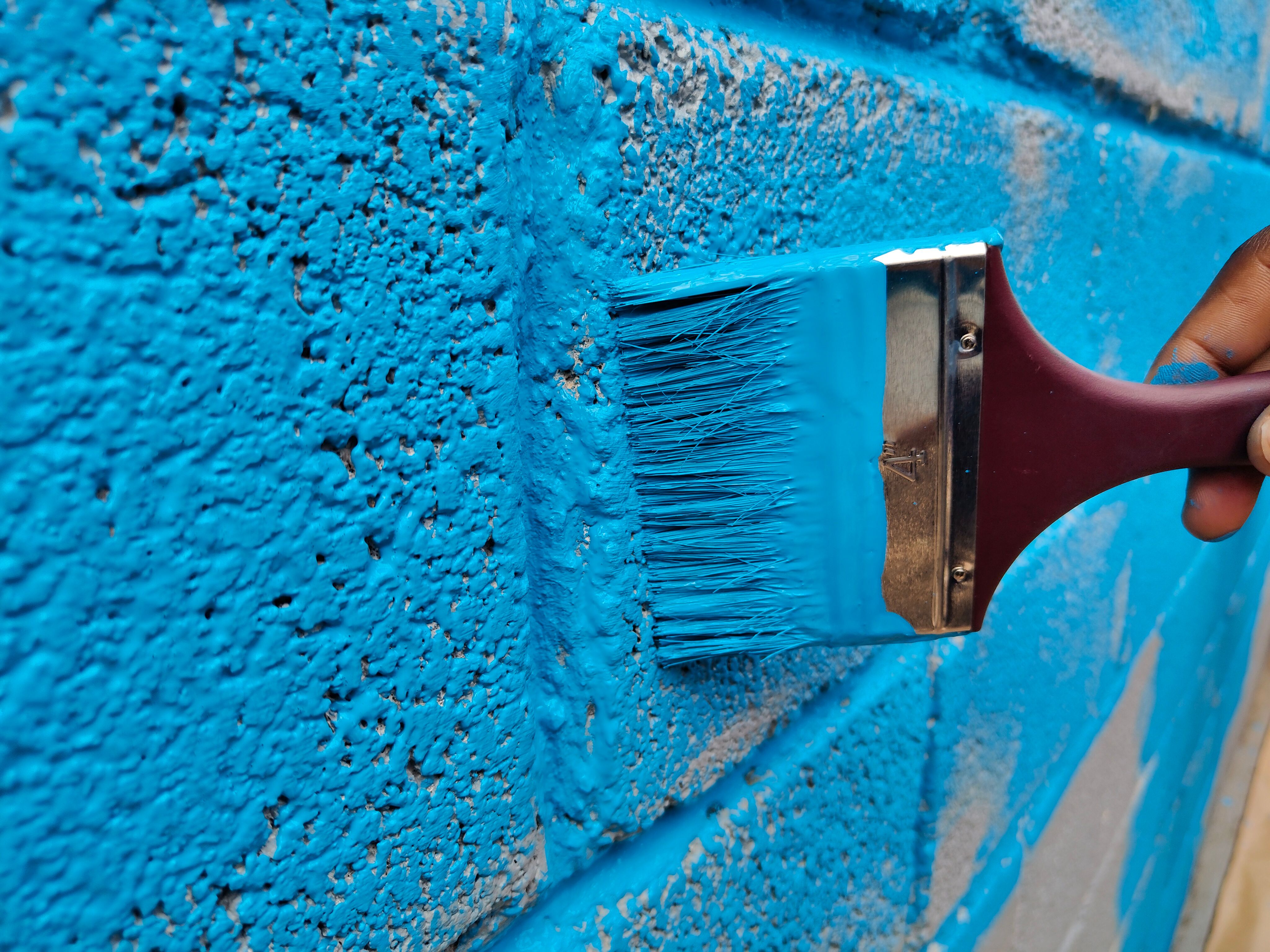 A hand painting a brick wall with vibrant blue paint using a brush.