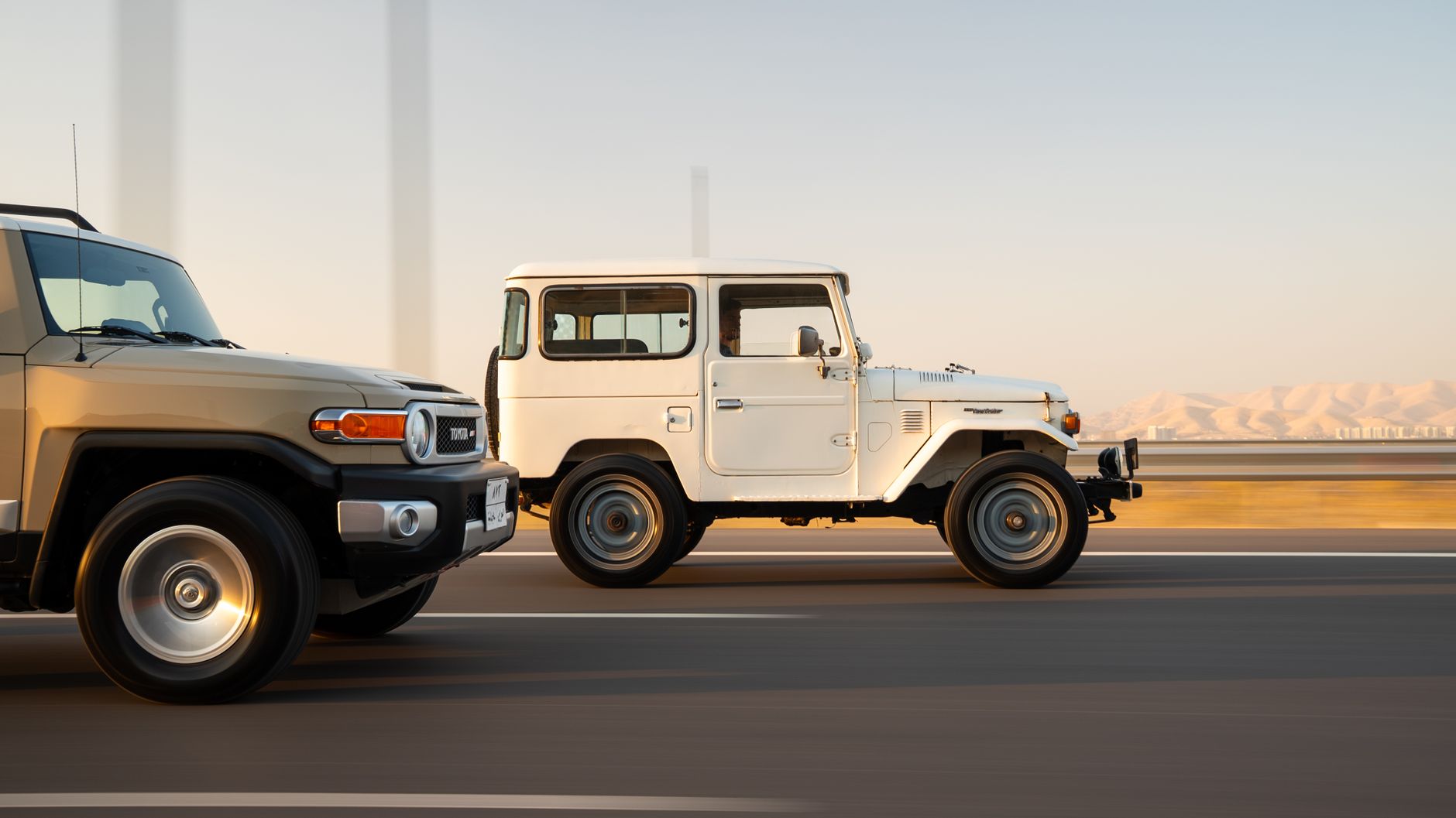 A vintage and modern SUV drive side by side on a scenic highway with mountains in the background.