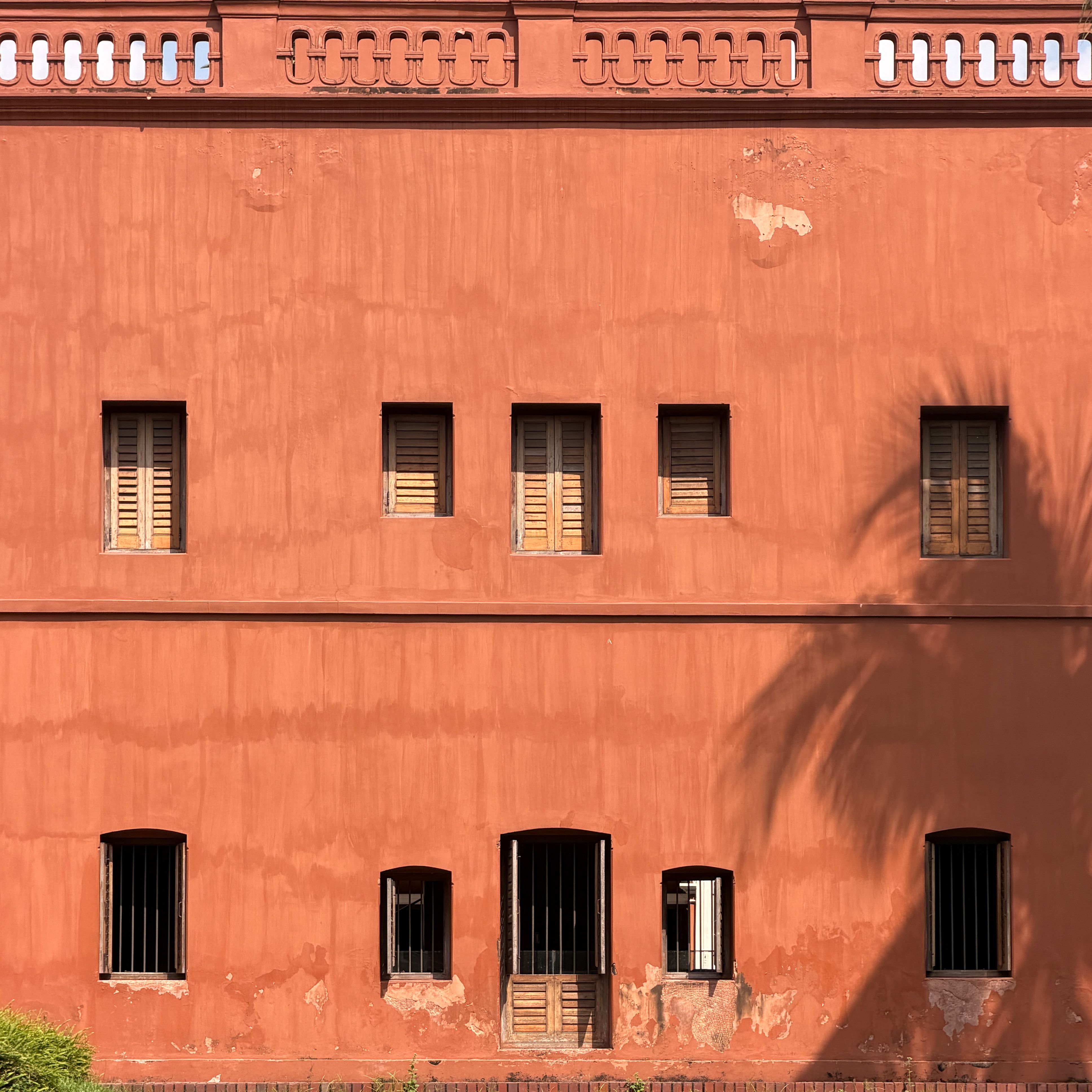 A historic red building facade with wooden shutters and shadow of a palm tree.