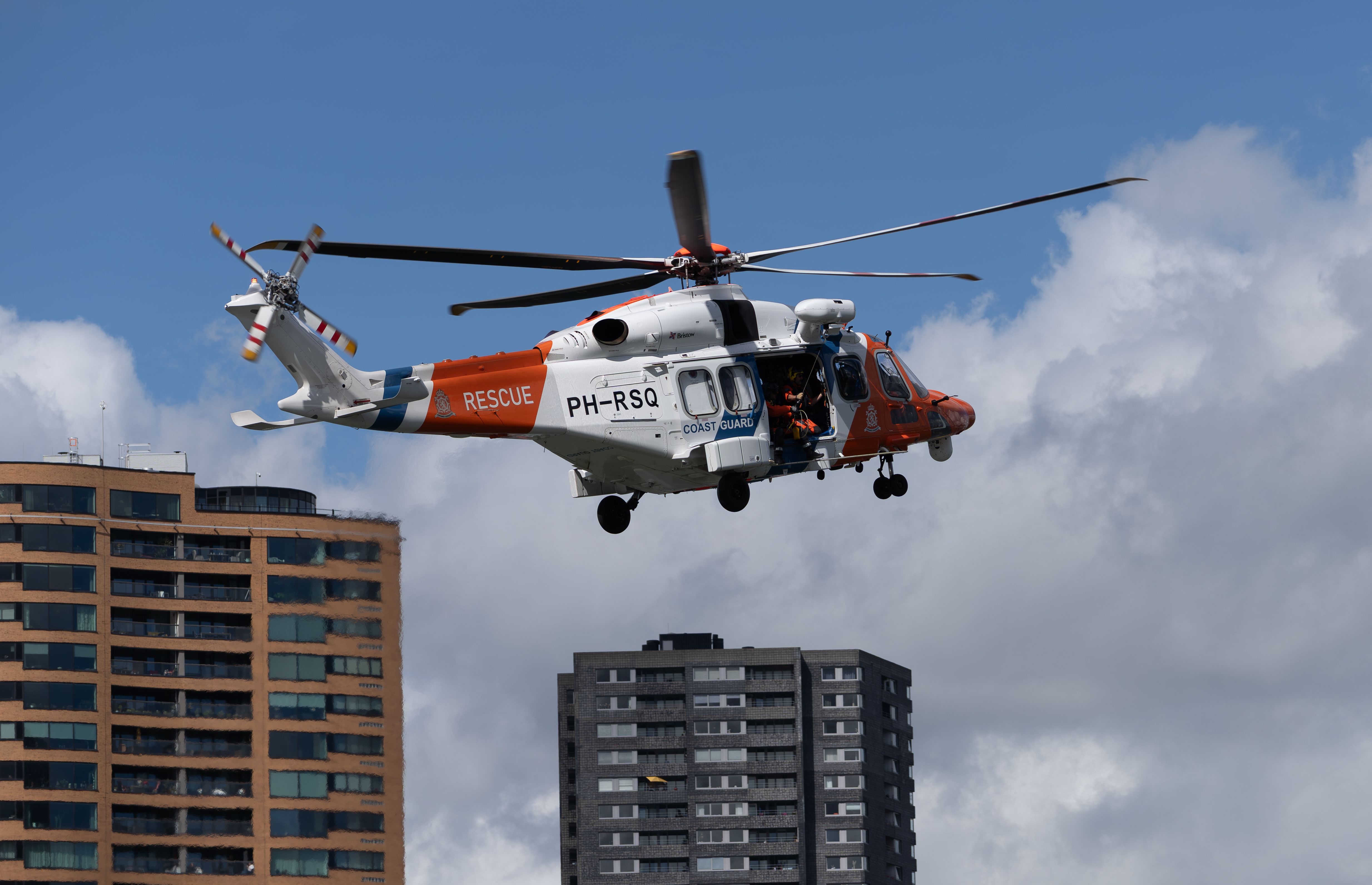 A coast guard rescue helicopter hovers over city buildings against a bright sky.
