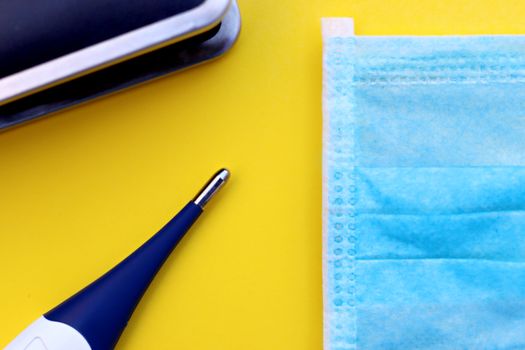 Flat lay of surgical mask and thermometer on yellow background, symbolizing pandemic protection.