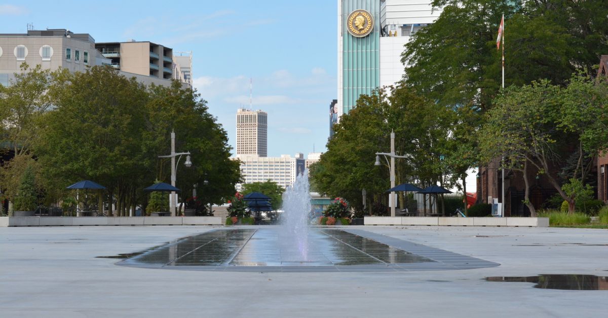 A tranquil city plaza with a central fountain and lush greenery under a clear blue sky.
