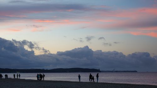 Free Silhouettes of people walking along a beach under a vibrant sunset sky with clouds. Stock Photo