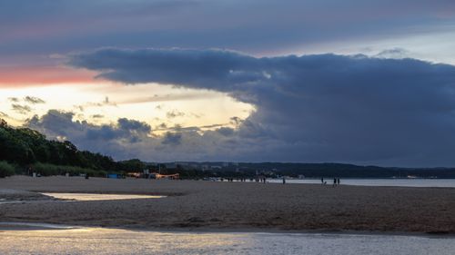 Free Calm beach scene with people at dusk, featuring colorful clouds and a tranquil shoreline. Stock Photo
