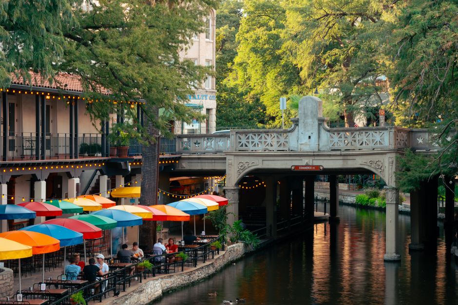 Colorful outdoor dining along the San Antonio Riverwalk with lush greenery and iconic bridge.