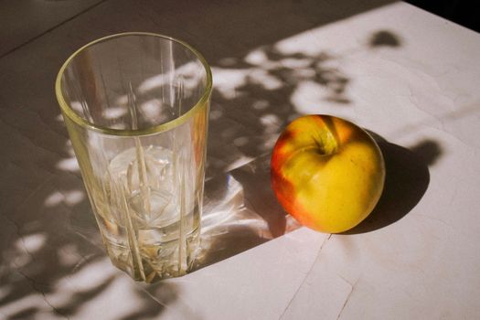 Minimalist still life featuring an apple and glass with sunlight and shadows on a table.