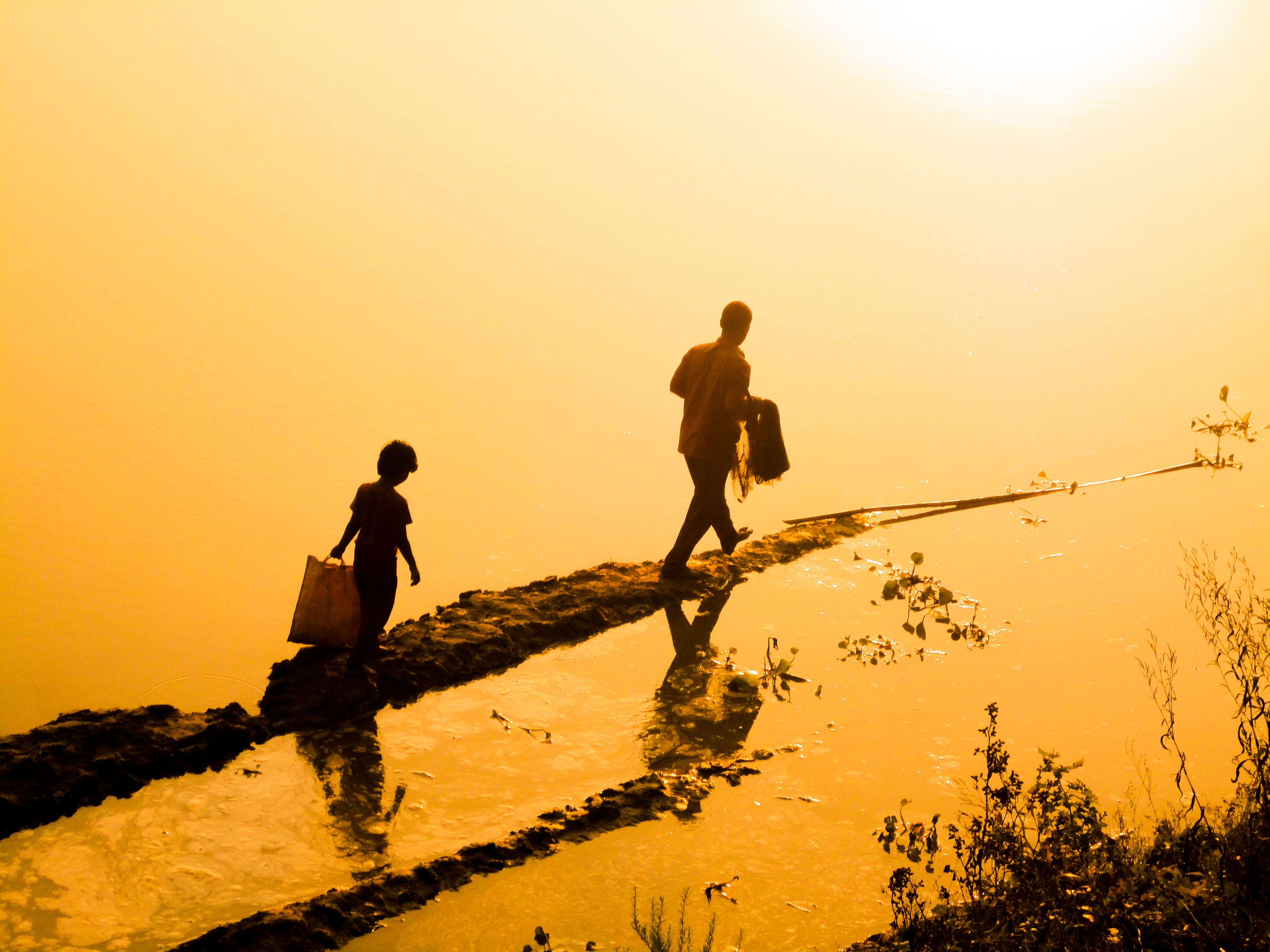 Silhouetted child and adult walking along a reflective path at sunrise, creating a serene and warm scene.