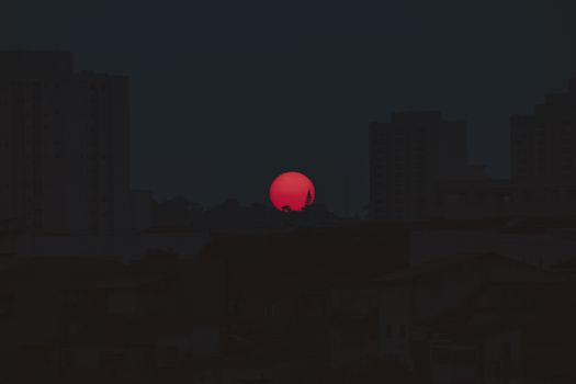 A vibrant red sun sets behind a silhouetted cityscape creating a dramatic evening scene.