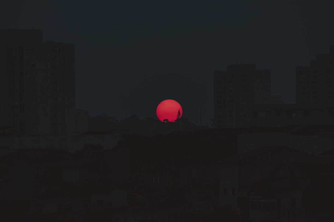 a vibrant red sun sets behind a silhouetted cityscape creating a drama