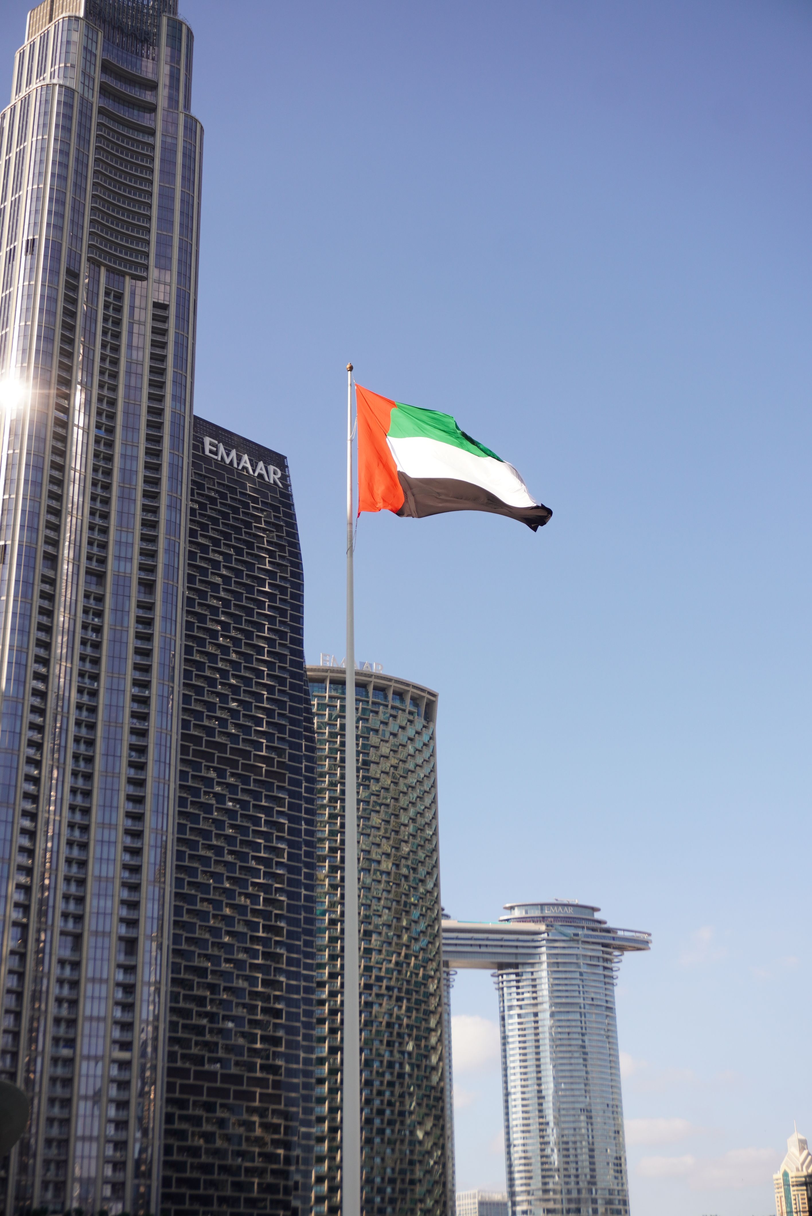 United Arab Emirates flag waving with Dubai skyscrapers in the background under a clear blue sky, showcasing modern architecture.