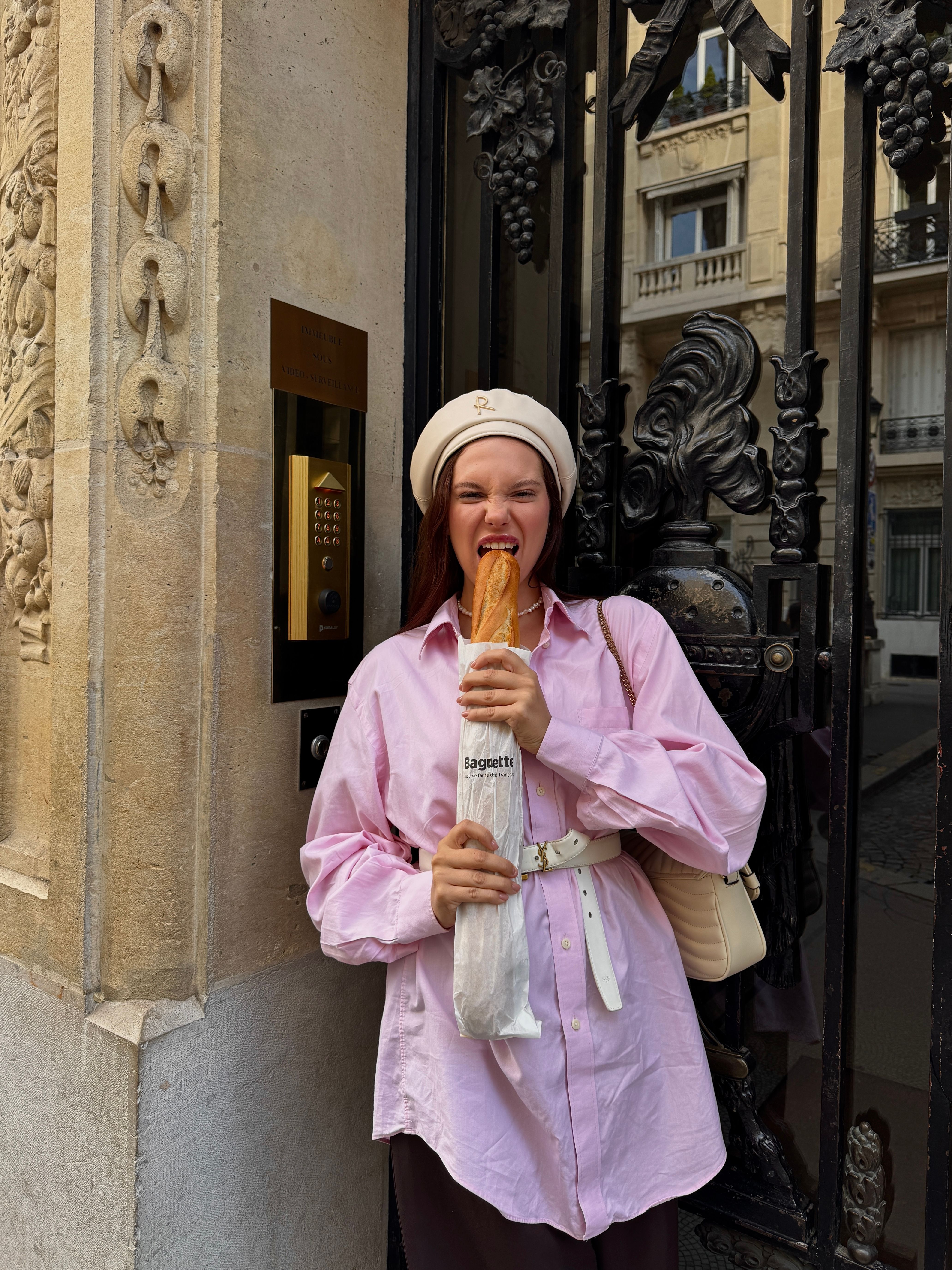 Smiling woman in a pink shirt holding a baguette in a French street setting.