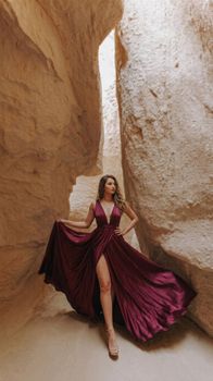 Woman in a flowing burgundy dress elegantly posed in a rocky cavern. Stunning natural setting.