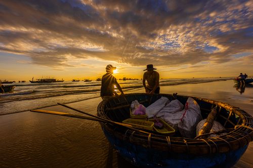 Free Silhouetted fishermen working on a beach at sunrise with boats and a vibrant sky. Stock Photo