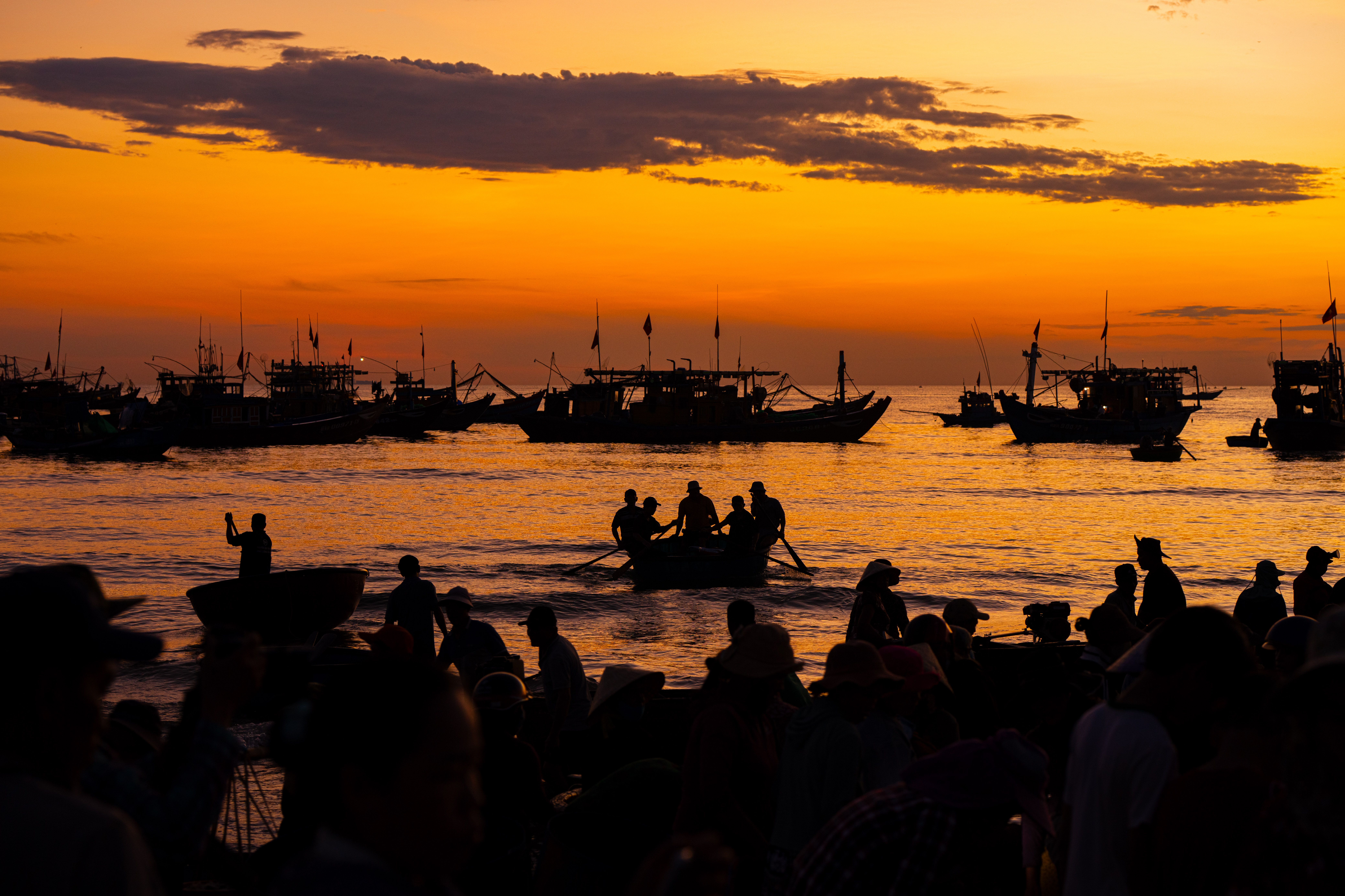 Silhouetted fishermen and boats against a vibrant orange sunset on the sea.