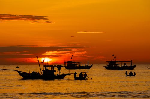 Free Silhouettes of fishing boats at sunset on calm ocean waters with vibrant orange sky. Stock Photo