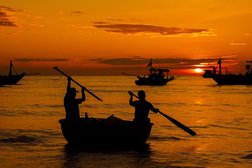Free A pair of silhouetted fishermen row their boat at sunrise on a tranquil sea backdrop. Stock Photo