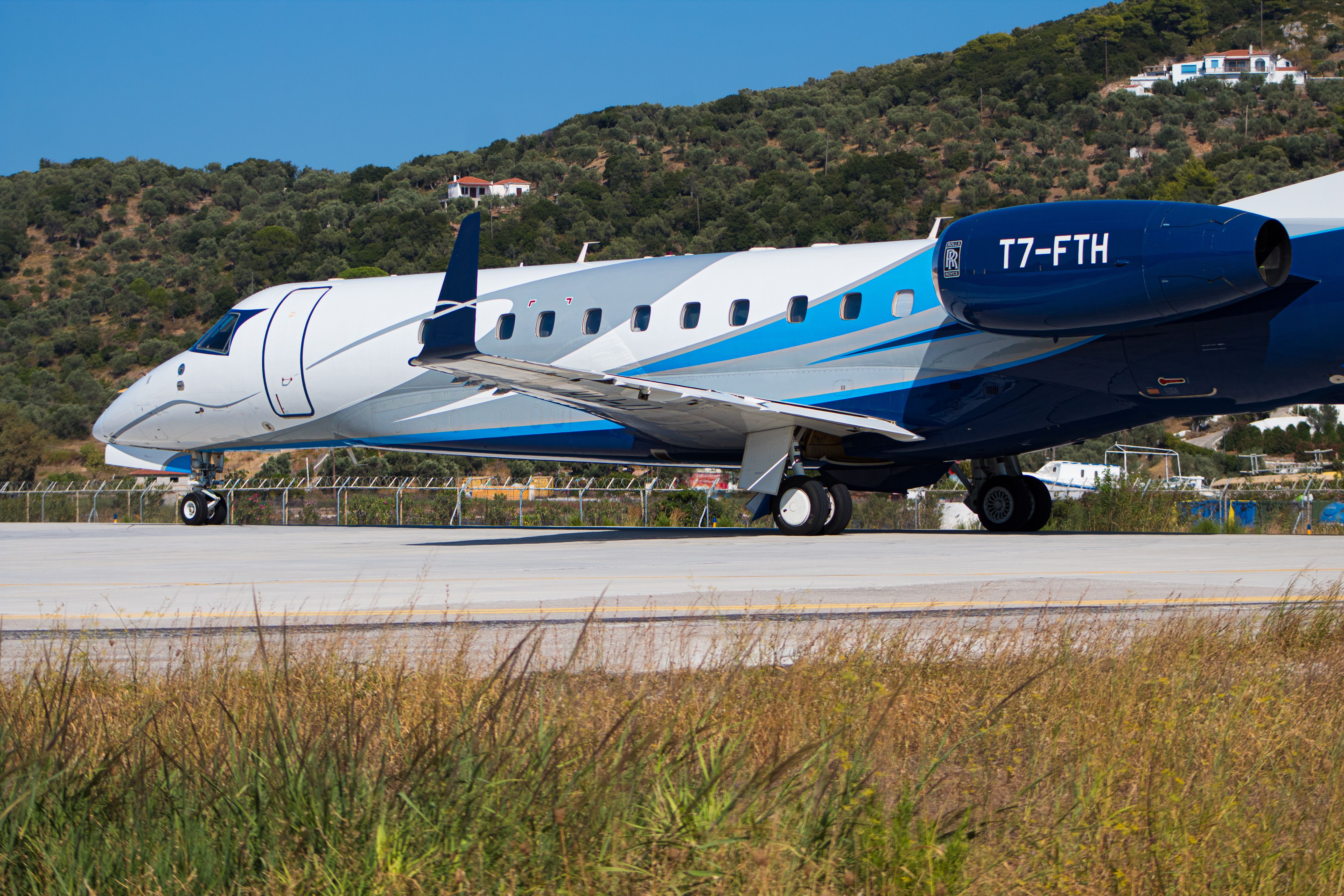 A sleek private jet positioned on a runway with a scenic Greek hillside backdrop.