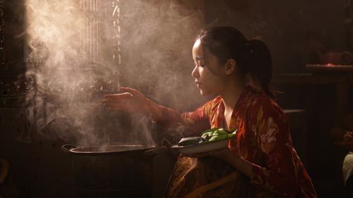 Free Indonesian woman in traditional attire cooking in a rustic kitchen with steam rising from a pot. Stock Photo