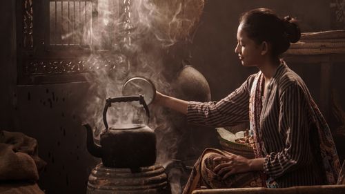 Free An Indonesian woman in traditional attire prepares tea on a clay stove in a rustic, smoky setting. Stock Photo
