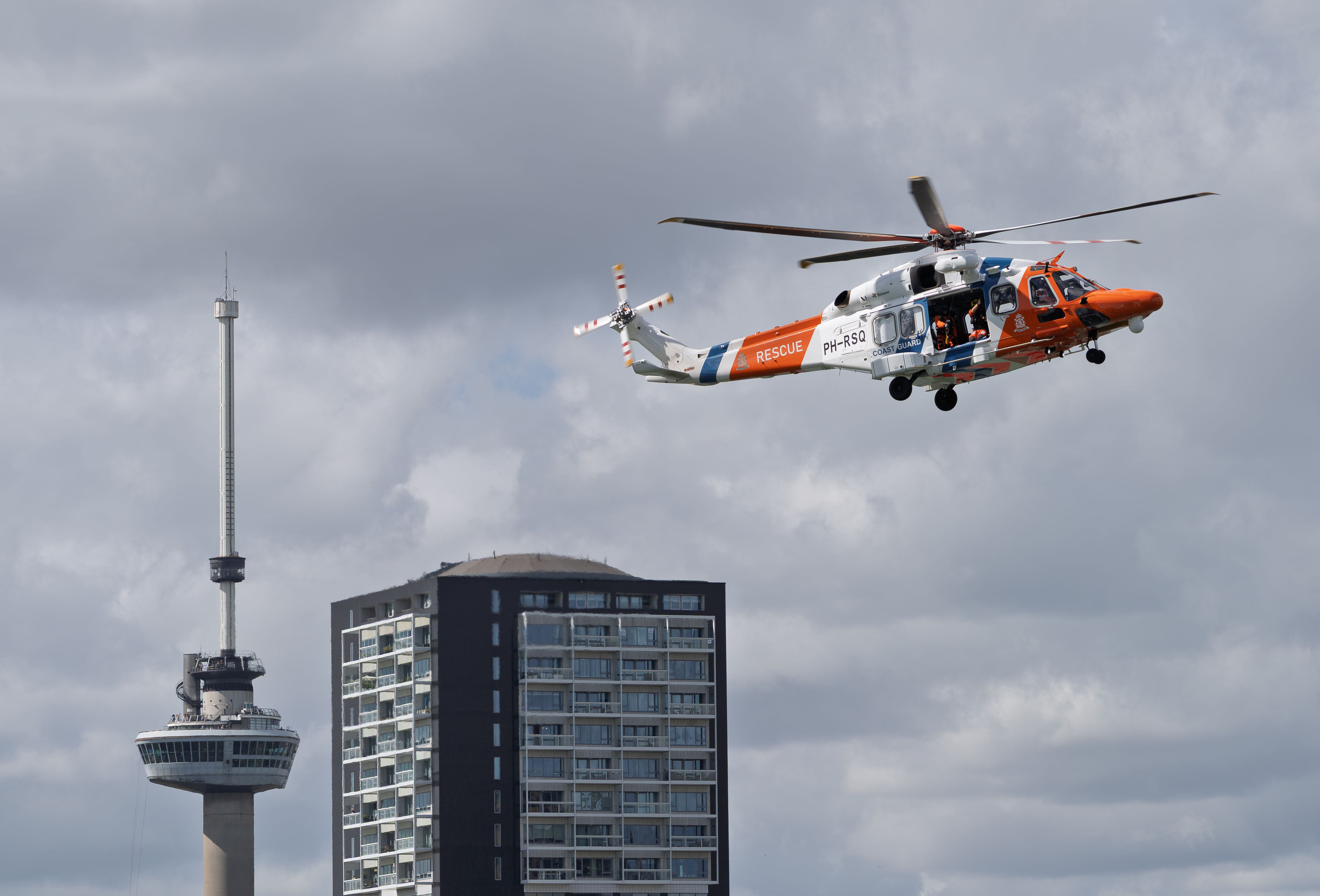 Rescue helicopter flying over Rotterdam cityscape with Euromast tower and apartment building.
