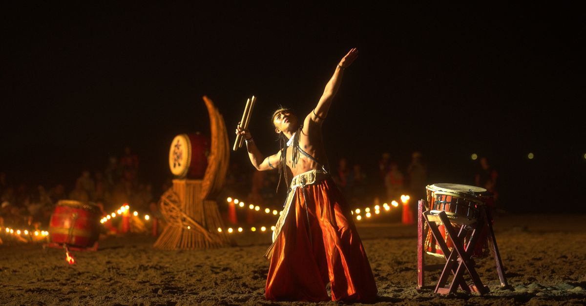 A taiko drummer energetically performs at a night event, surrounded by ambient lighting and traditional instruments.