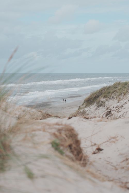 Free Two people walking on a serene beach surrounded by sandy dunes and ocean waves. Stock Photo